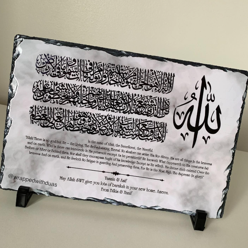 A rectangular slate wall plaque featuring black Arabic calligraphy and English translation text, displayed on two small black stands against a light beige wall.