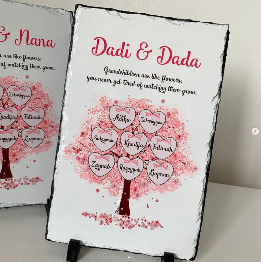 Personalized rectangular slate sign with black slate edges, featuring a pink and red family tree design with heart-shaped names, displayed on a small stand against a light wall.