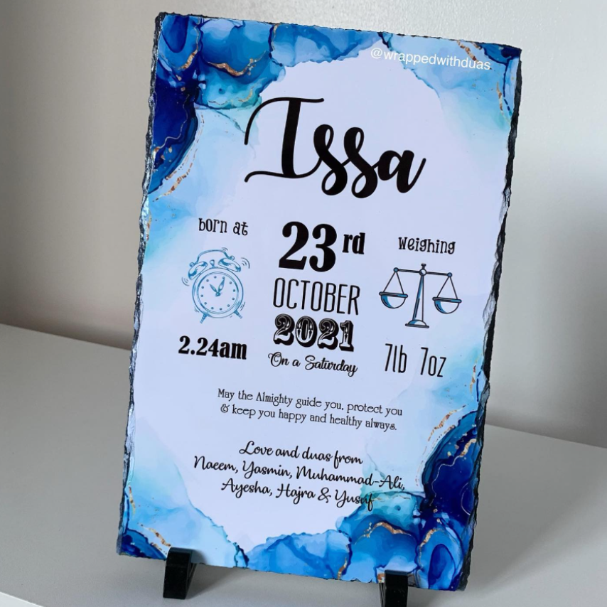 A personalized slate birth announcement plaque with a blue and white marbled design, displayed on a small black stand against a neutral gray background.