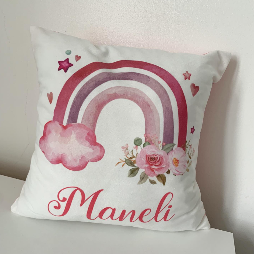 A square white throw pillow with a watercolor-style pink and purple rainbow, pink flowers, small hearts, and the name 'Maneli' in pink script, resting on a white surface against a light wall.