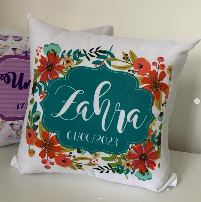 A square white throw pillow with a colorful floral border and a central teal label reading 'Zahra 01/06/2023' in white script, photographed on a light surface against a beige wall.