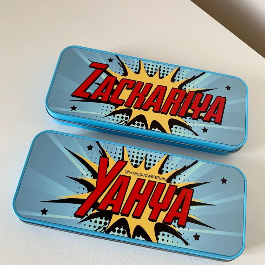 Two blue metal pencil cases with comic-style burst graphics and large red text reading 'Zachariya' and 'Yahya' on a white surface.