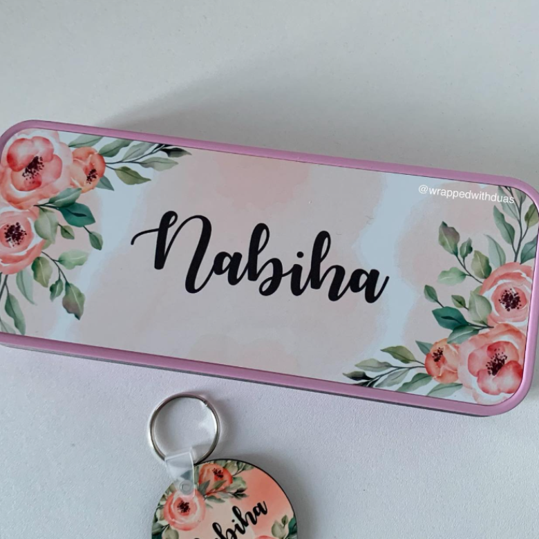 A rectangular metal tin with a pink border and floral watercolor design in coral, pink, and green, personalized with the name 'Nabiha' in black script. A matching round keychain tag with a silver key ring is placed below the tin on a light gray surface.