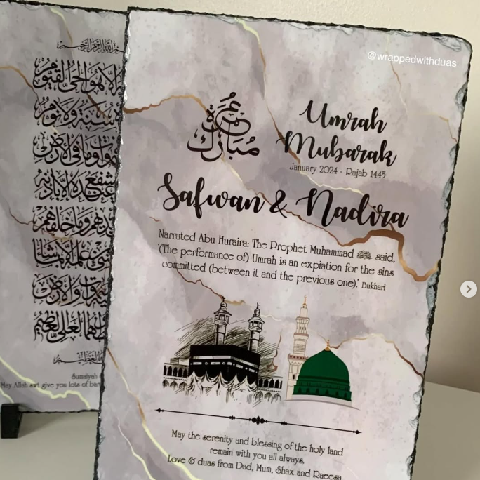 Personalized Islamic wedding anniversary slate plaque with 'Umrah Mubarak' and 'Salwan & Nadira' text, mosque illustration, and gold veining on a white marble-look surface, displayed on small black stands.