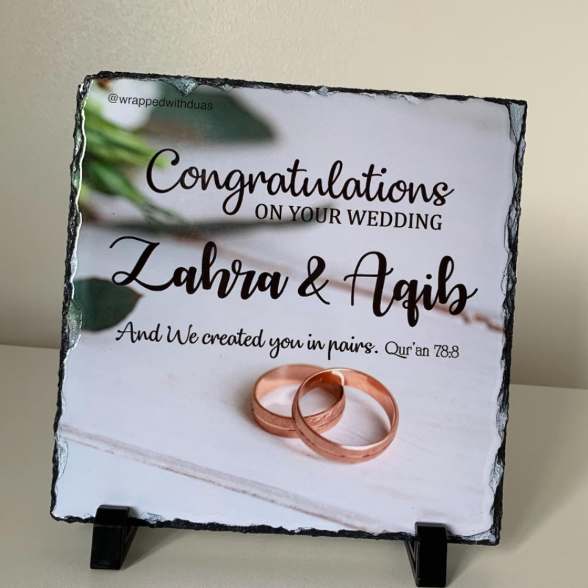 A square slate wedding sign on a small black display stand, featuring the text 'Congratulations on your wedding Zahra & Aqib' with two rose-gold rings printed on a white background. The sign is photographed indoors against a light beige wall and surface.