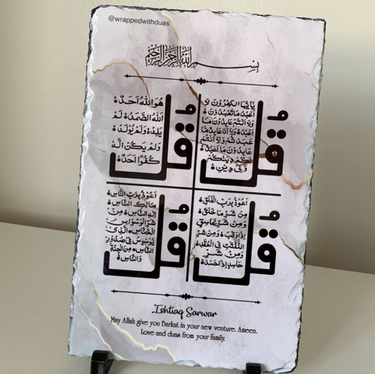 A rectangular slate plaque with a white marbled surface, featuring large black Arabic calligraphy and smaller Arabic text, plus the English phrase 'Ishiq Sawar' and a blessing at the bottom. The plaque is displayed on small black stands against a light beige wall.