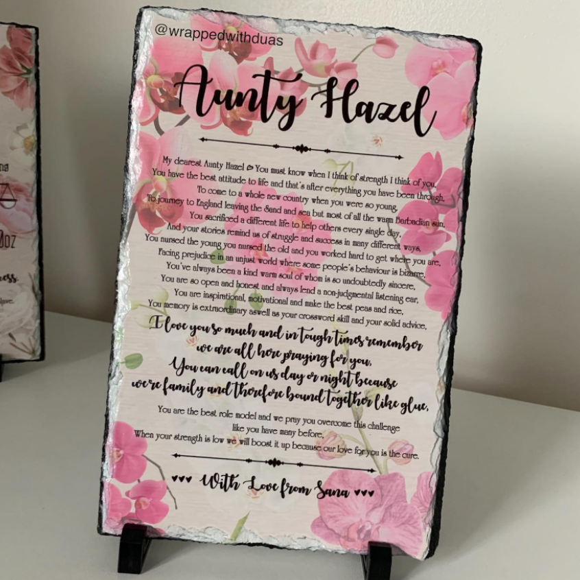A rectangular black slate sign with rounded corners, printed with a pink floral border and the handwritten name 'Aunty Hazel' above a sentimental poem, photographed leaning against a wall.