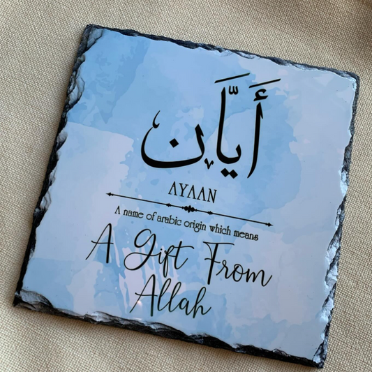 A square slate gift plaque with a light blue watercolor background, featuring the Arabic word 'Ayat' in black calligraphy, the English name 'AYAAN', a small explanatory phrase, and 'A Gift From Allah' in script. The plaque is photographed on a beige textured fabric background.