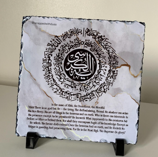 A square decorative slate plaque with black edges and natural marbling, featuring a central black circular calligraphy motif and Arabic text. The lower portion has additional printed text in English, and the plaque is displayed on a small black stand against a light beige wall.