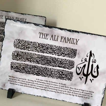 A decorative black-and-white slate plaque on a small stand, printed with the text 'THE ALI FAMILY,' Arabic calligraphy, and a short English quote. Another similar plaque is partially visible behind it against a light beige wall.