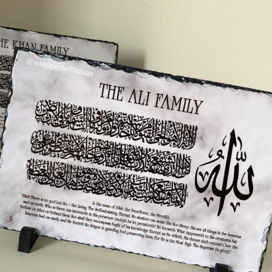 A decorative black-and-white slate plaque on a small stand, printed with the text 'THE ALI FAMILY,' Arabic calligraphy, and a short English quote. Another similar plaque is partially visible behind it against a light beige wall.