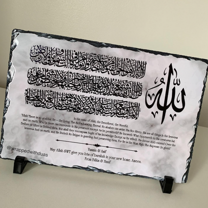 A rectangular slate wall plaque featuring black Arabic calligraphy and English translation text, displayed on two small black stands against a light beige wall.