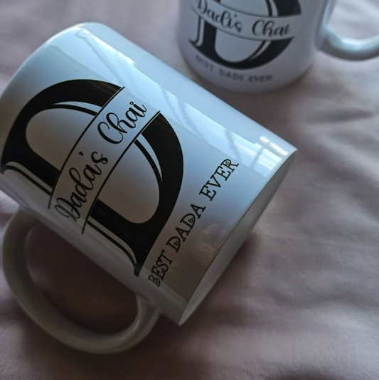 Two white ceramic mugs printed with a large black letter D, the words 'Dad's Chai,' and 'Best Dada Ever,' resting on a soft pink fabric surface.