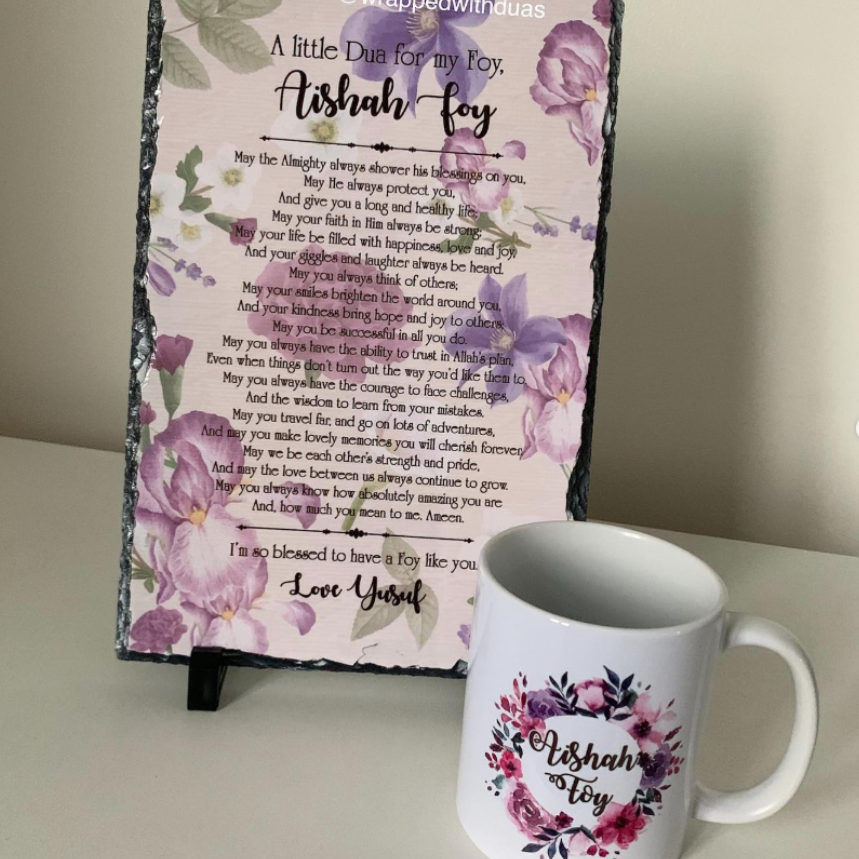 A floral printed sign with a poem titled 'A Little Due for My Foxy Aishah-fey' stands on a small black easel, next to a white ceramic mug with a matching floral 'Aishah-Fox' design. The scene is photographed indoors against a light wall.