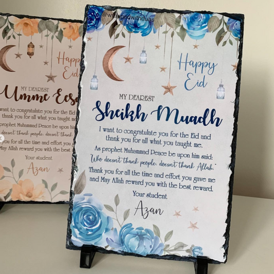 A personalized slate sign for Eid Mubarak featuring blue floral illustrations, hanging lanterns, crescent moons, and printed congratulatory text, displayed on a small stand.