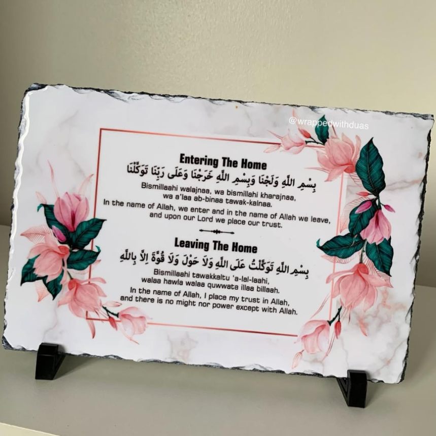 A rectangular slate sign with irregular dark edges, printed with floral artwork in pink and green and bilingual text about entering and leaving the home, displayed on two small black stands against a beige wall.
