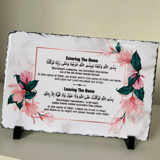 A rectangular slate sign with irregular dark edges, printed with floral artwork in pink and green and bilingual text about entering and leaving the home, displayed on two small black stands against a beige wall.