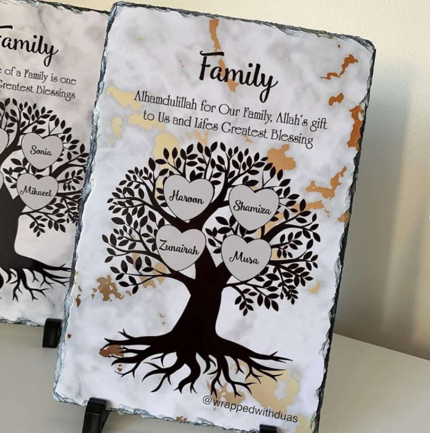 Two personalized slate family tree signs displayed on black stands against a light beige wall. The front sign features a white marbled background with black slate edges, a black tree silhouette, and five heart-shaped branches holding the names of family members.