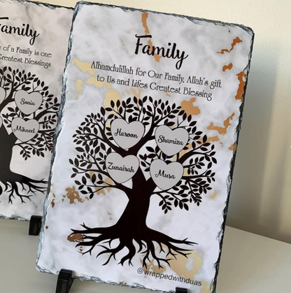 Two personalized slate family tree signs displayed on black stands against a light beige wall. The front sign features a white marbled background with black slate edges, a black tree silhouette, and five heart-shaped branches holding the names of family members.