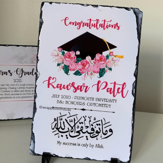 A personalized graduation sign with a white background, black slate-style edges, pink script text, a black graduation cap illustration, and pink floral artwork. The sign is displayed upright on a small black stand against a light beige wall.