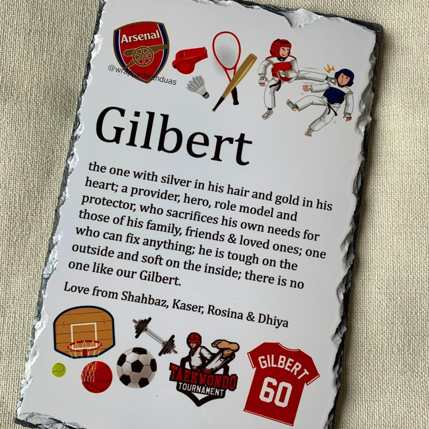 A personalized black slate plaque featuring a custom sports-themed memory quote for 'Gilbert' with various sports logos and illustrations, photographed on a beige textured fabric background.