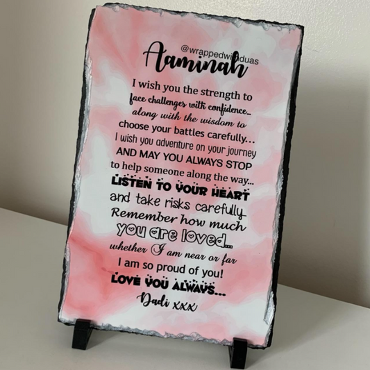 A personalized rectangular slate sign with a pink watercolor background and black text, displayed on a small black stand against a neutral beige wall.