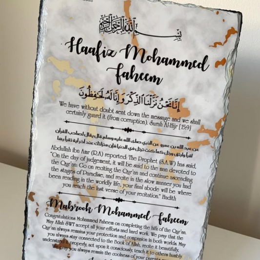 A rectangular slate plaque on a small black display stand, featuring printed Islamic calligraphy and English text honoring Prophet Muhammad. The plaque has a white background with brown distressed edges and gold-toned decorative accents.