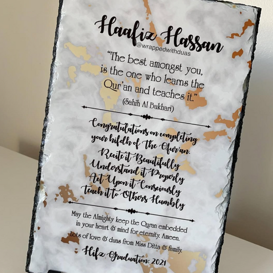 A square slate sign on a small black display stand, featuring a white marble-style background with gold abstract marbling and black text celebrating a Hafiz graduation with a Quran quote.