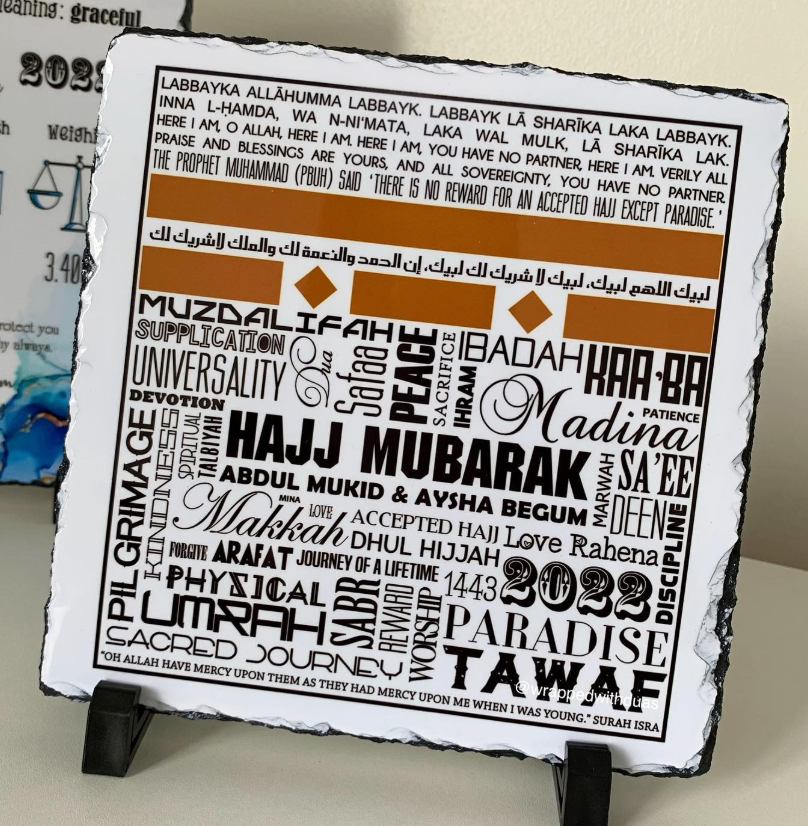 A square white slate or stone plaque with black edges, featuring Arabic calligraphy and a typographic design in brown and orange text, displayed on a small black stand against a light background.