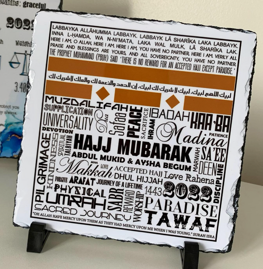 A square white slate or stone plaque with black edges, featuring Arabic calligraphy and a typographic design in brown and orange text, displayed on a small black stand against a light background.