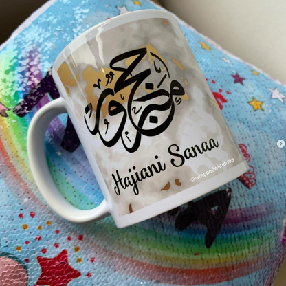 A white ceramic mug with black Arabic calligraphy and the text 'Hajjani Sanaa' resting on a colorful, star-patterned zippered pouch.
