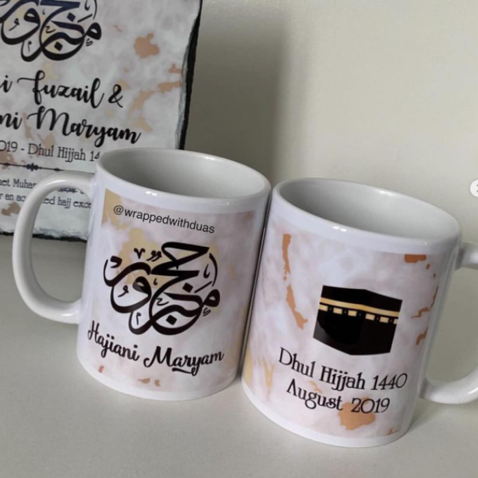 Two personalized white ceramic mugs with gold and black Islamic calligraphy and text, displayed on a light surface with a matching printed card in the background.