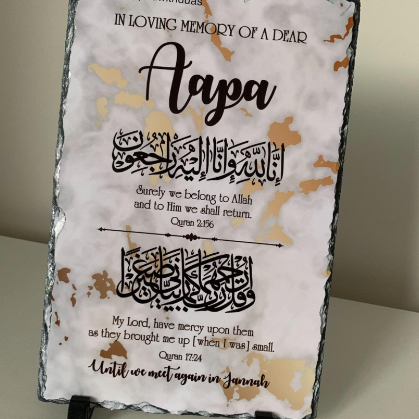 A rectangular memorial plaque with a distressed white and gold background, featuring black Arabic calligraphy and English remembrance text, displayed on a small stand against a beige wall.