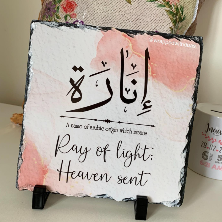 Square slate sign on a small black stand, featuring Arabic calligraphy and the English phrase 'Ray of light; Heaven sent' on a white background with soft pink watercolor washes. The sign is photographed indoors on a tabletop with decorative fabric and a mug in the background.