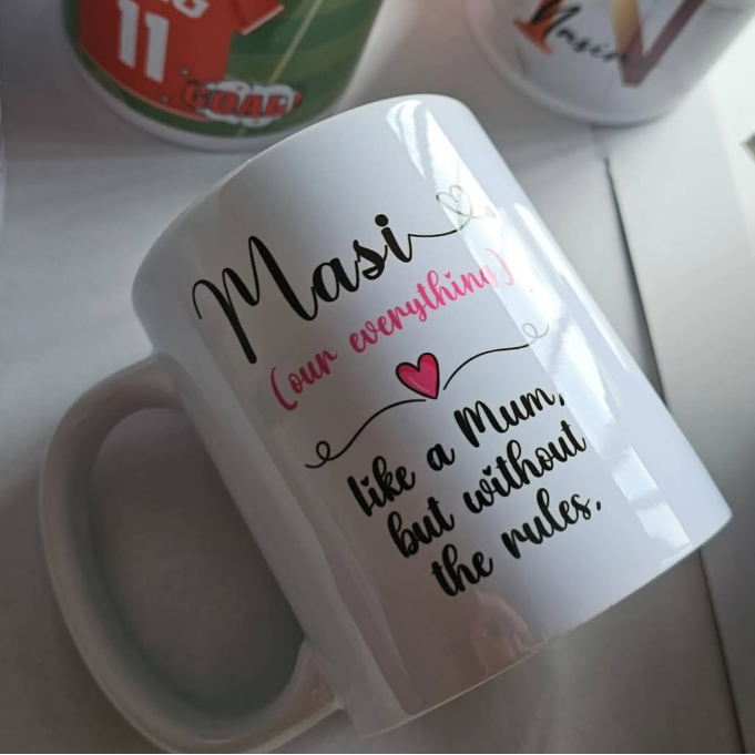 A white ceramic mug with a custom printed message reading 'Masi (our everything) like a Mum, but without the rules.' in black and pink text with a small pink heart, shown in the foreground with two other mugs blurred in the background.