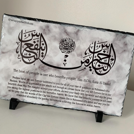 A rectangular slate plaque with black edges and a white marble-look surface, displayed on two black stands against a light beige wall. The plaque features black Arabic calligraphy, a central emblem, and a block of English text at the bottom.