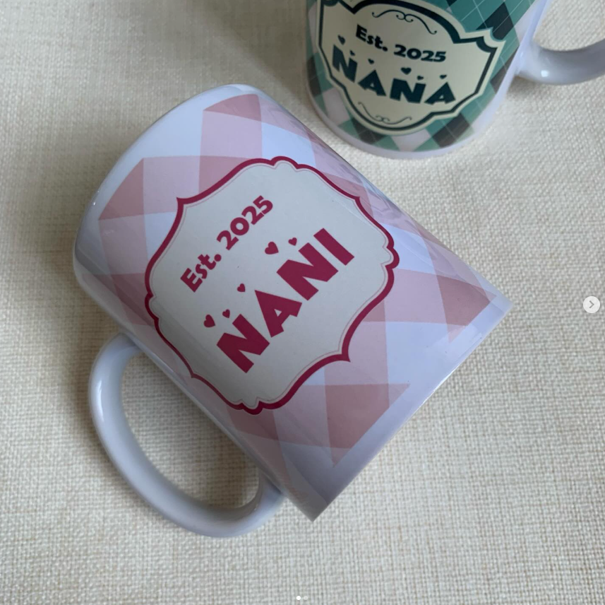 A white ceramic mug with a pink and white gingham pattern and a central red label reading 'Est. 2025 NANI' is shown on a beige fabric surface.