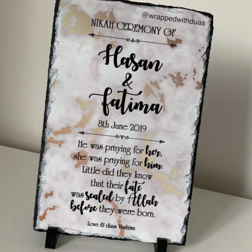 A personalized slate sign with black text reading 'Nikah Ceremony of Hasan & Fatima' and a poem, displayed on a small black stand against a light beige wall.