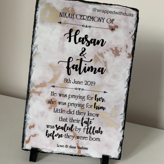 A personalized slate sign with black text reading 'Nikah Ceremony of Hasan & Fatima' and a poem, displayed on a small black stand against a light beige wall.