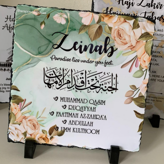 Square slate sign with a floral watercolor design in sage green, peach, and cream, featuring the text 'Zeinab' and Islamic verses. The sign is displayed on a small black stand in front of other decorative signs.