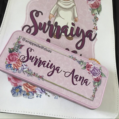 A pink rectangular metal case with floral illustrations and the personalized name 'Suraiya Anna' in purple script, placed on top of a matching printed pouch or notebook.