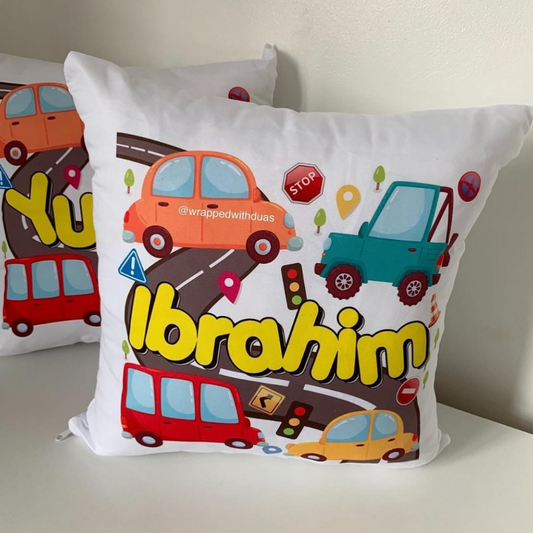 A white square throw pillow featuring a colorful cartoon road scene with multiple cars, traffic lights, and the name 'Ibrahim' in large yellow letters. A second similar pillow is partially visible behind it on a white surface against a light wall.