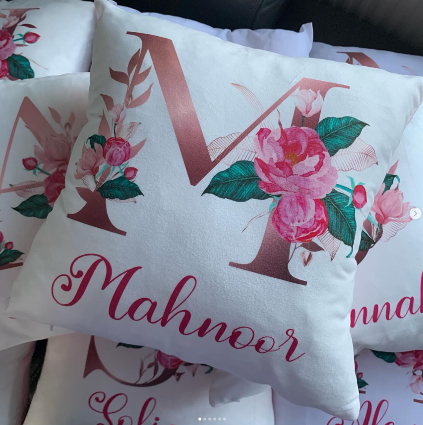 A square white pillow featuring a large metallic rose gold letter 'M' decorated with pink and green floral designs, and the name 'Mahnoor' in pink cursive script. Other matching pillows are partially visible in the background.