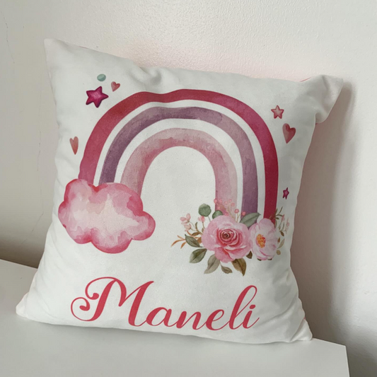 A square white throw pillow with a watercolor-style pink and purple rainbow, pink flowers, small hearts, and the name 'Maneli' in pink script, resting on a white surface against a light wall.