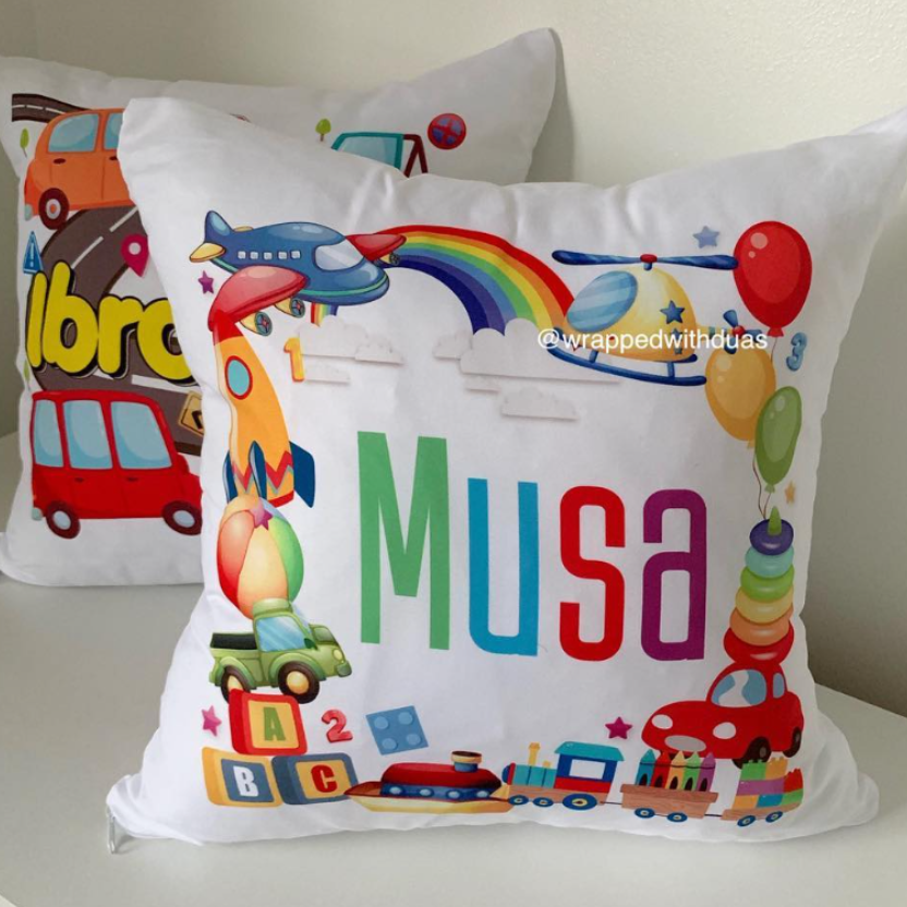Two personalized white throw pillows with colorful children's toy and rainbow graphics, featuring the name 'Musa' on the front pillow, displayed on a white shelf against a beige wall.