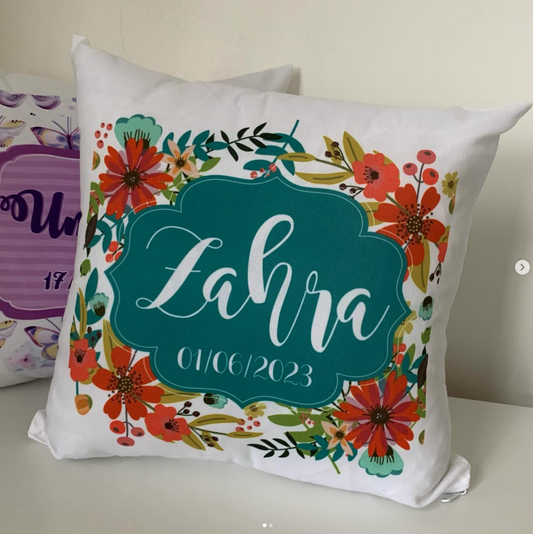 A square white throw pillow with a colorful floral border and a central teal label reading 'Zahra 01/06/2023' in white script, photographed on a light surface against a beige wall.