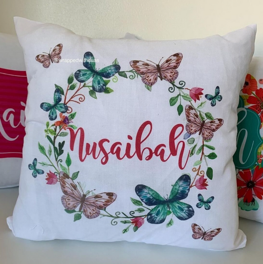A square white throw pillow featuring a personalized design with the name 'Nusaibah' in red script, surrounded by a colorful floral and butterfly wreath. The pillow is displayed on a white surface with other decorative pillows partially visible in the background.