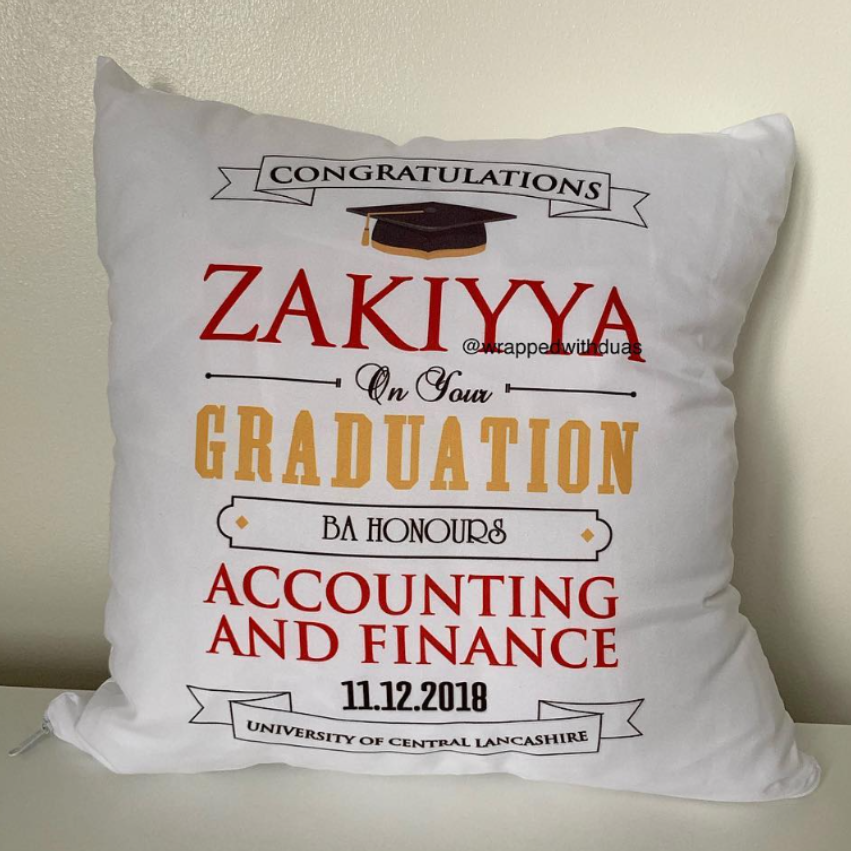 A white square graduation throw pillow with personalized printed text and graphics, leaning against a beige wall on a light surface.
