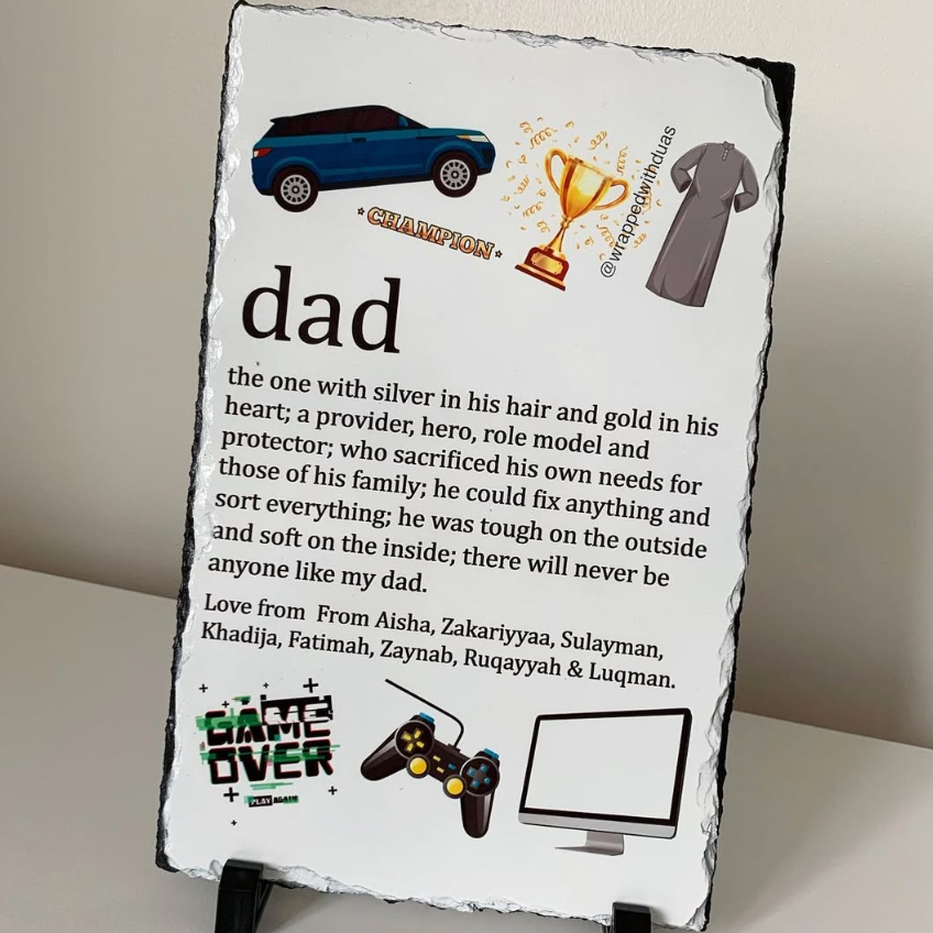 A rectangular slate sign with a black natural stone border and a white front panel, displayed standing upright against a light beige wall. The panel features a printed 'dad' definition with small illustrated graphics including a blue car, trophy, robe, game controller, and computer monitor, plus a 'Game Over' graphic and a 'Happy Dad' message.