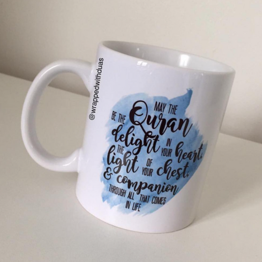 A white ceramic mug with a blue watercolor-style graphic and black script text reading 'May the Quran be the delight in your heart, the light of your chest, and companion through all that comes in life.' The mug is photographed on a light beige surface against a neutral beige background.